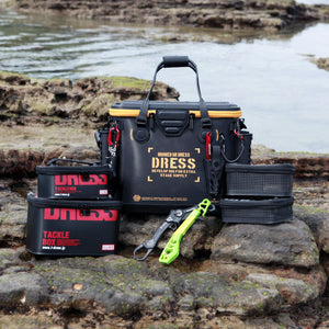 A black DRESS Tackle Supply cooler bag and matching storage cases are arranged on a rocky shoreline with water in the background. The main bag is black with yellow accents and printed DRESS branding, with two smaller black cases, a green folding tool, and a black mesh basket placed in front.
