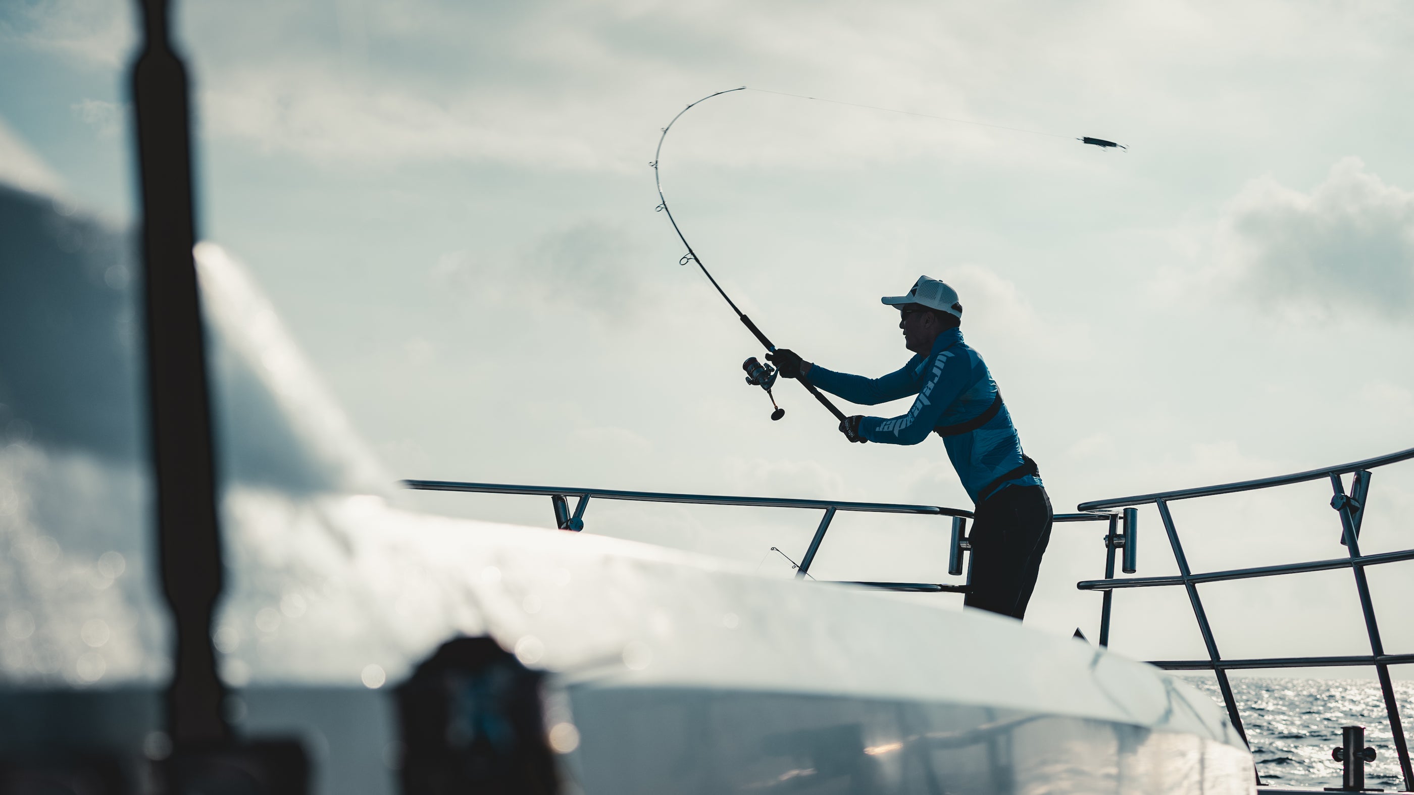A person stands on a boat deck holding a bent lureleader fishing rod, silhouetted against a bright sky with clouds and water in the background.