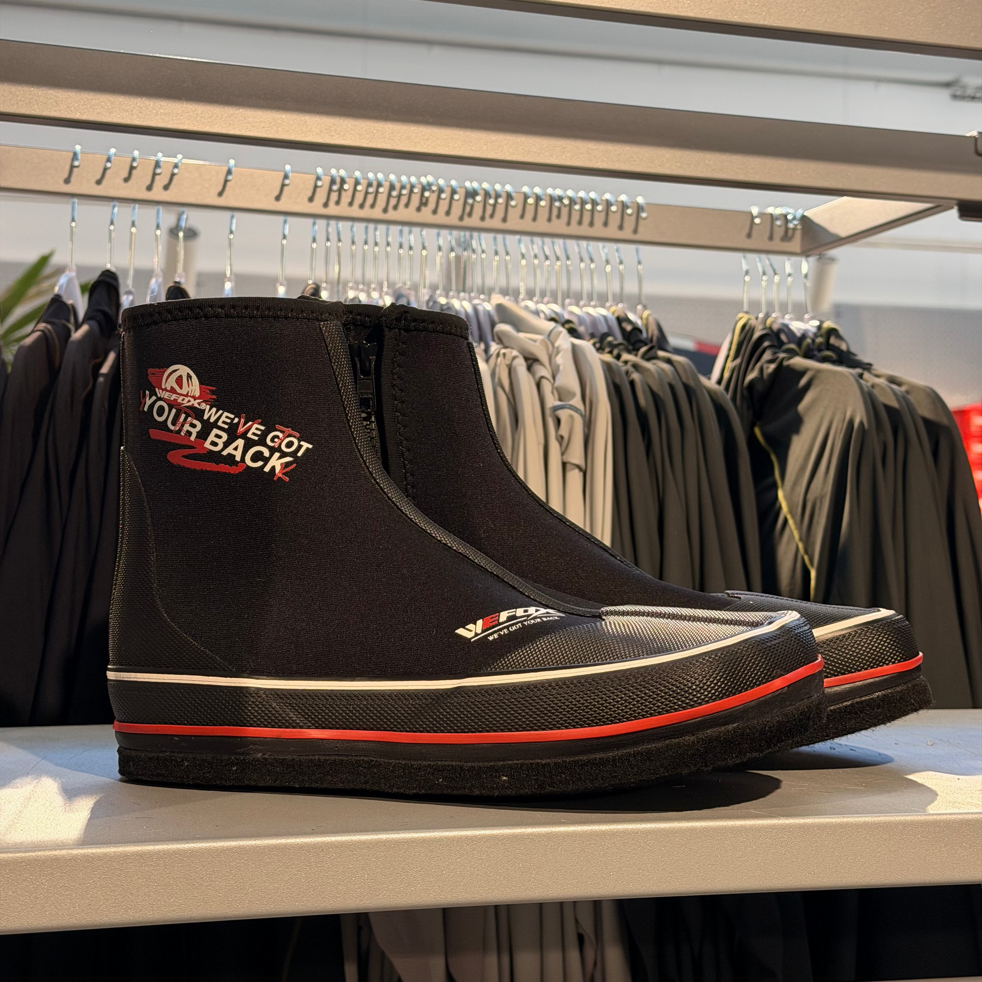A pair of black mid-calf waterproof boots from Wefox with red and white accents, displayed on a retail shelf with clothing racks in the background.