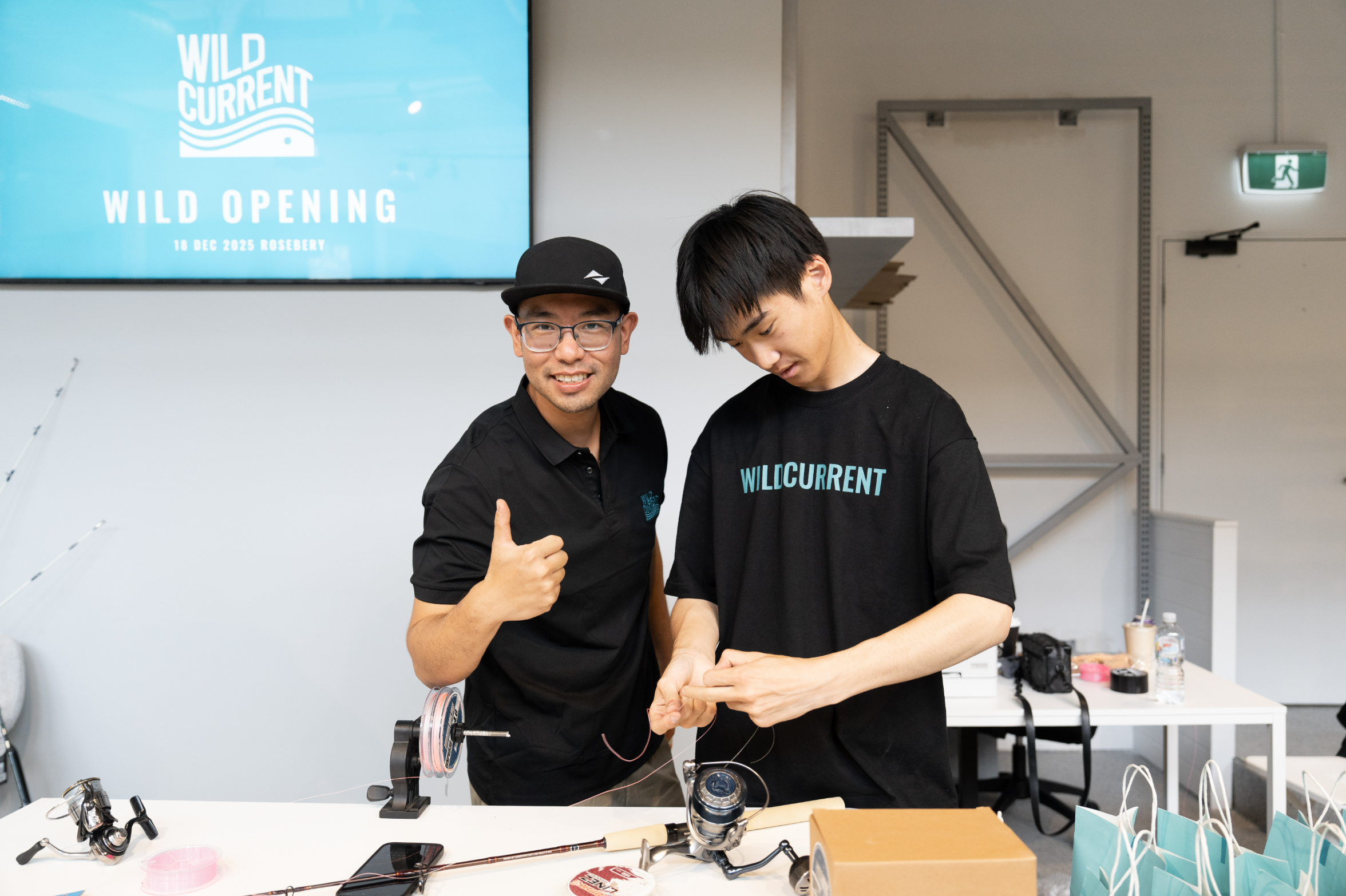 Two men standing in a bright office or studio space, both wearing black shirts with the brand name 'WILD CURRENT' printed on the chest. One man gives a thumbs-up while wearing a black cap, and the other looks down at his hands.