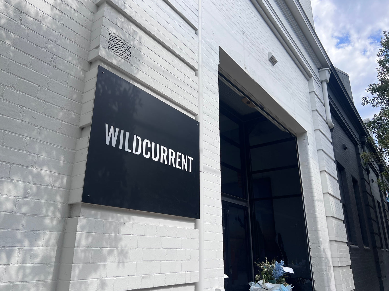 A black rectangular sign with the brand name 'WILDCURRENT' in white uppercase letters, mounted on a white painted brick building facade next to a dark doorway.